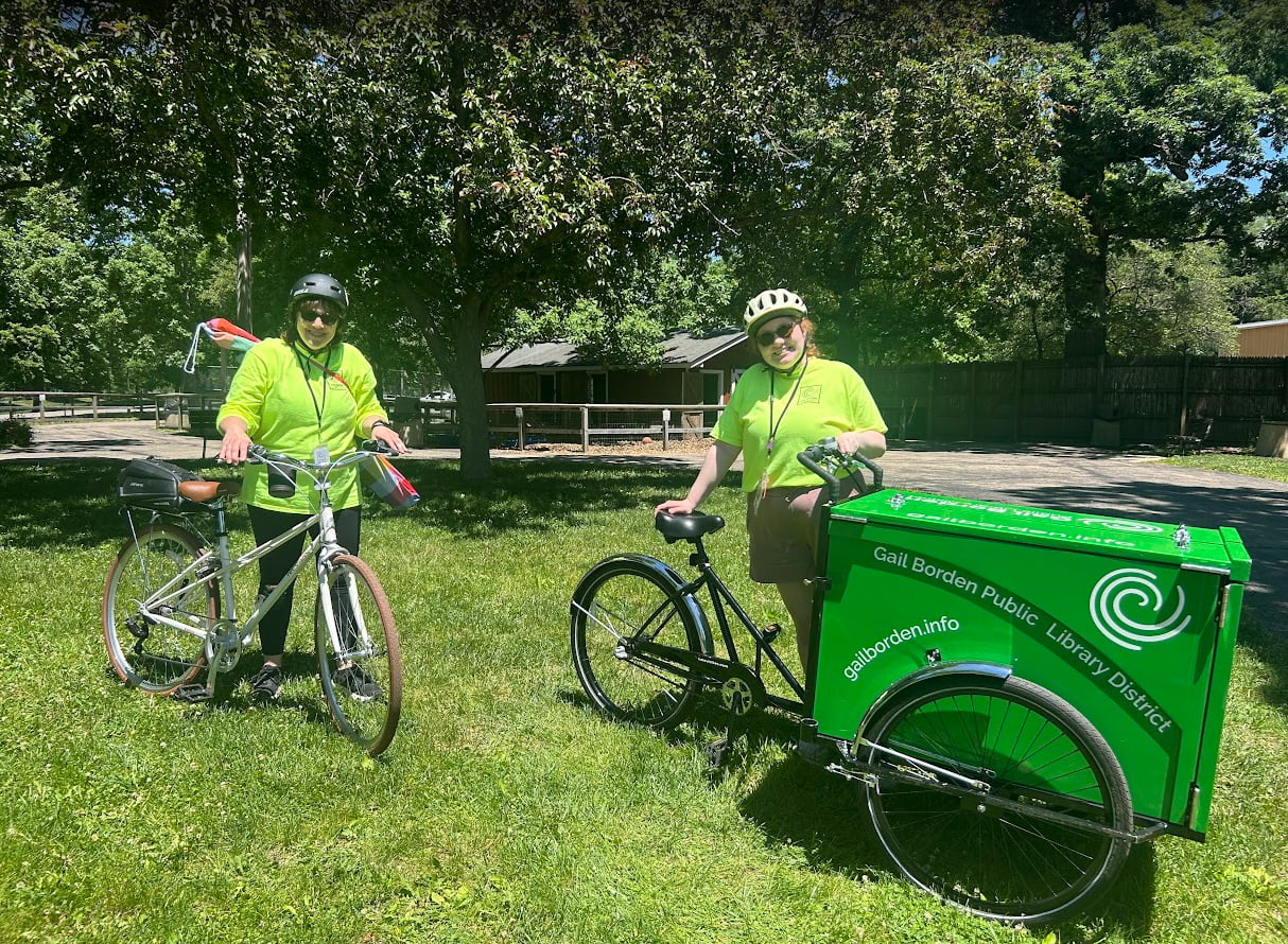 Two staff members wearing hi vis shirts and helmets stand next to their bicycles.  One is a standard bicycle, and the other is a tricycle with a large green crate between the two front wheels.