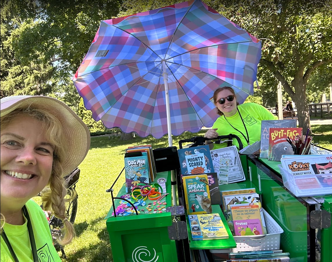 The crate on the tricycle is opened up, revealing shelves and bins full of books.  A large parasol shades the staff members and the bikes from the sun.