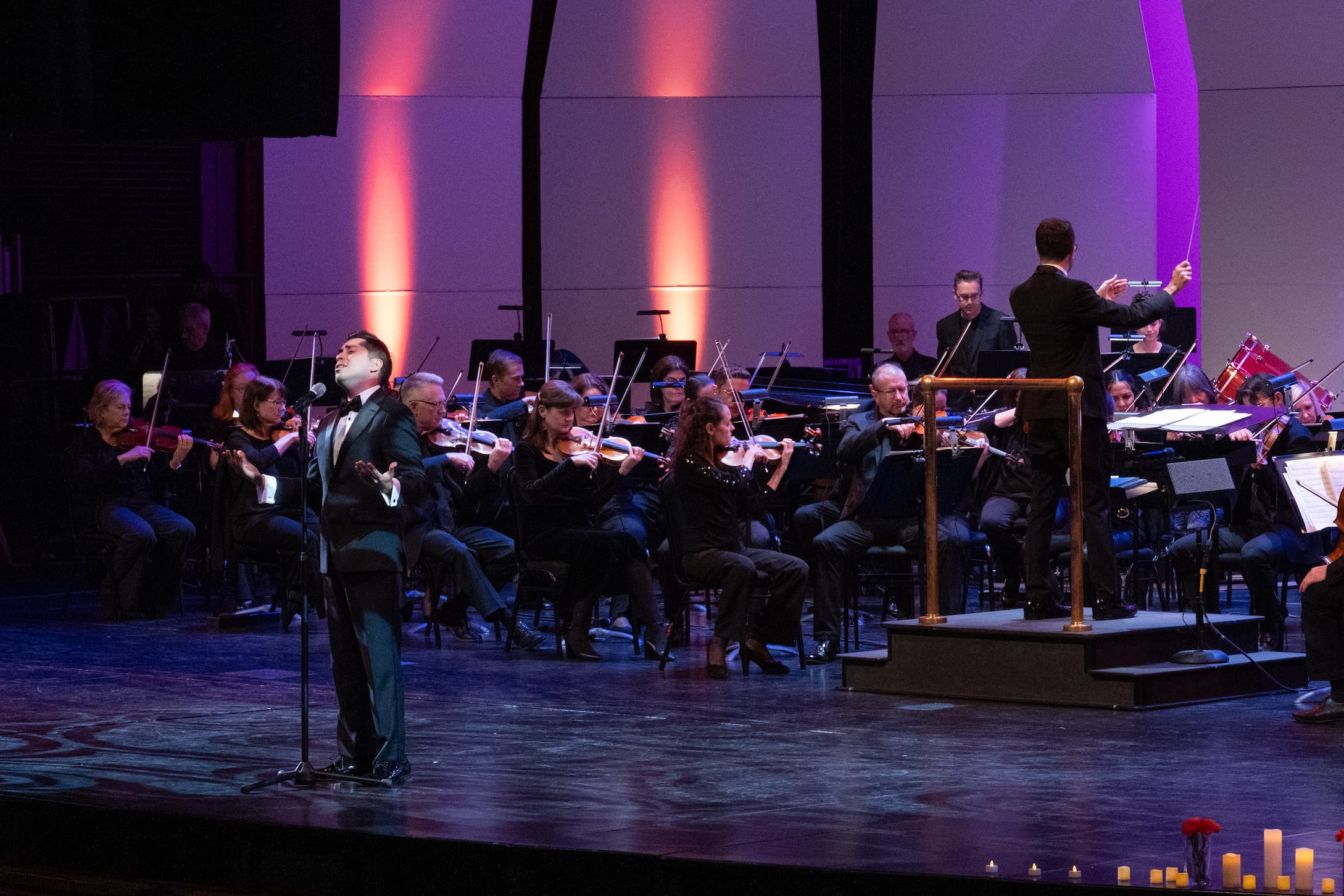 A vocalist in a tuxedo performs on stage in front of the Elgin Symphony Orchestra during the Día de los Muertos concert, celebrating cultural traditions through music and performance.