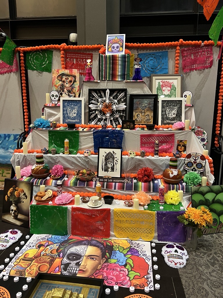 A community ofrenda at the Hemmens Culture Center features framed artwork, candles, papel picado, and flowers arranged together to honor and remember loved ones during Día de los Muertos.