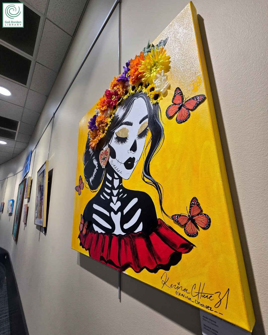 Artwork inspired by Día de los Muertos traditions, displayed in a library gallery. The painting depicts a stylized skeletal figure adorned with a colorful floral crown and butterflies against a vibrant yellow background.