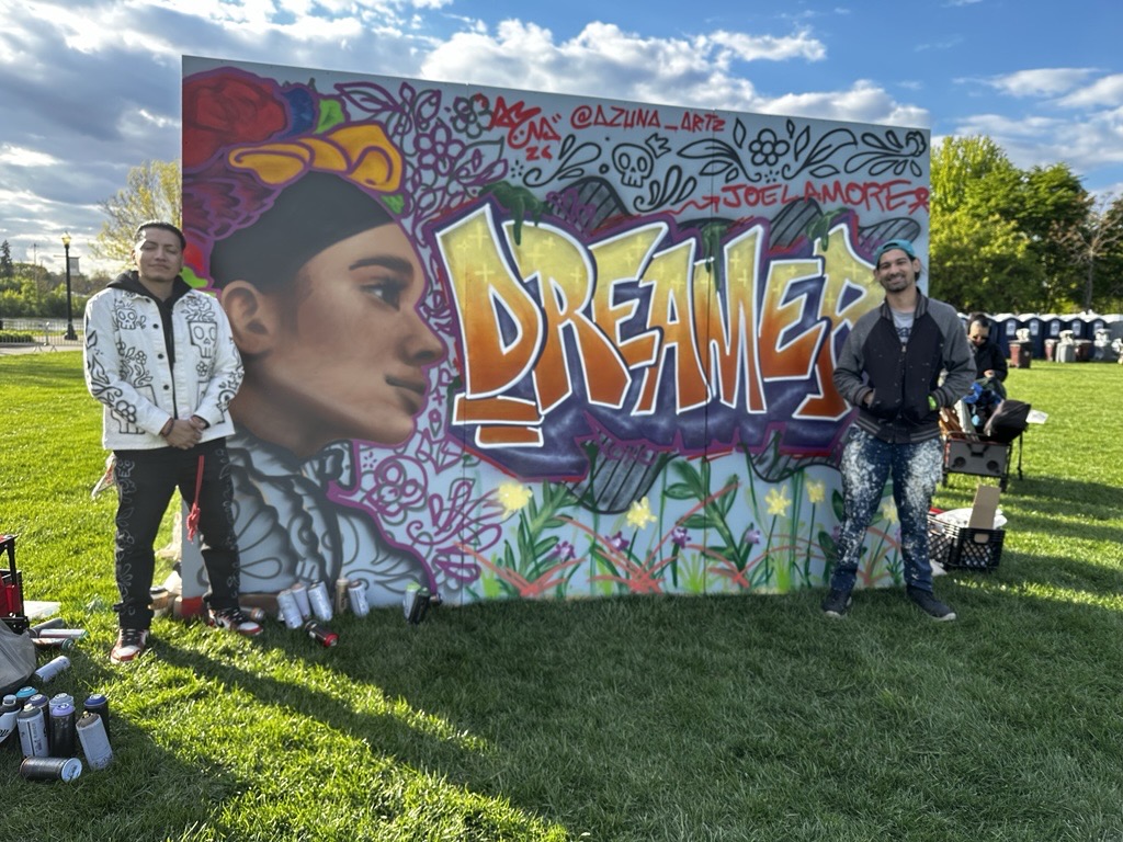 Outdoor mural featuring a stylized portrait and the word “Dreamer” in graffiti-style lettering. The artwork incorporates floral elements and vibrant colors, while two artists stand on either side of the completed mural.