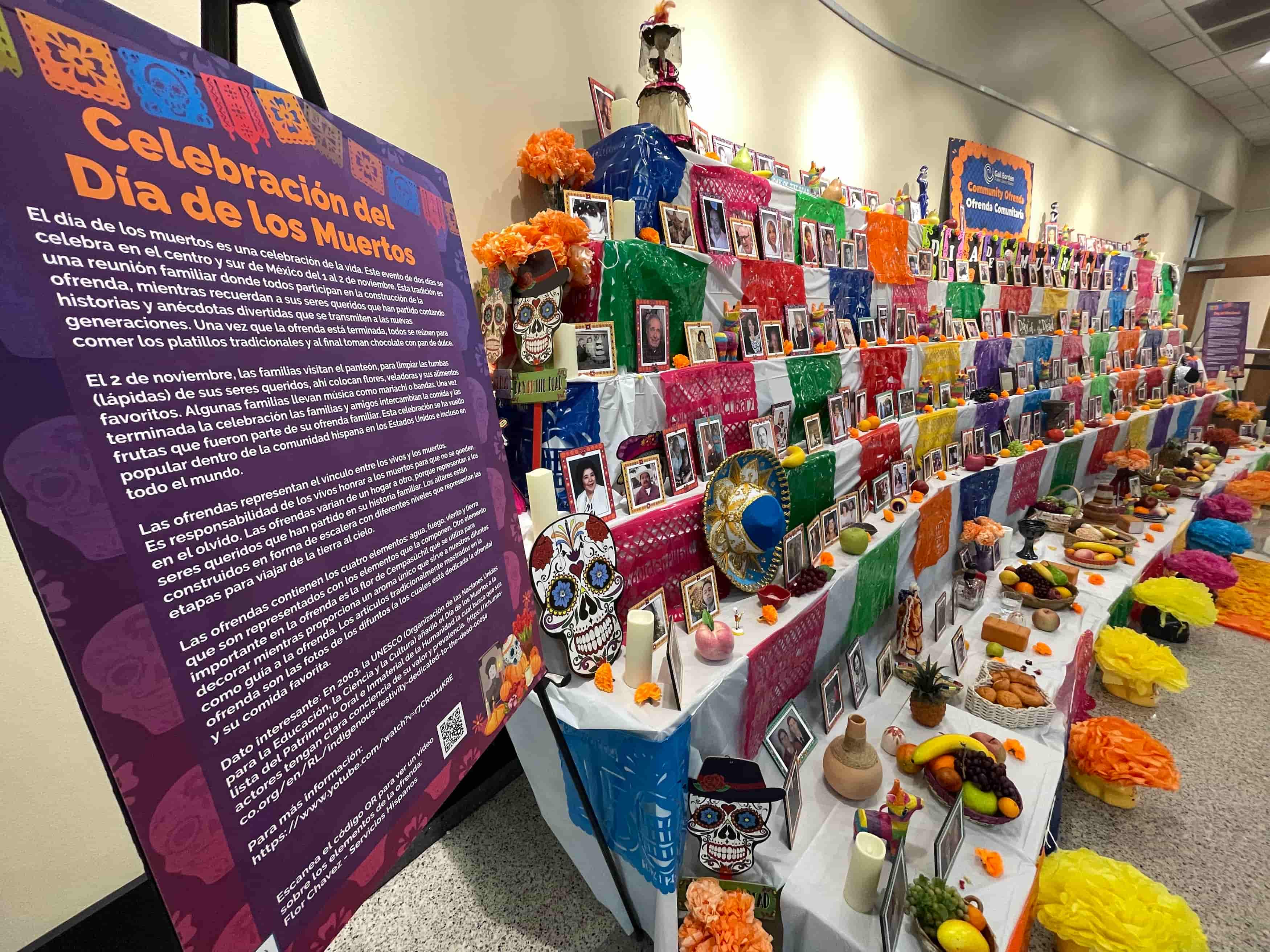 Community Día de los Muertos Ofrenda displayed in the library, decorated with papel picado, flowers, commemorative photographs, and traditional foods. The installation honors and remembers loved ones through elements representative of this cultural tradition.